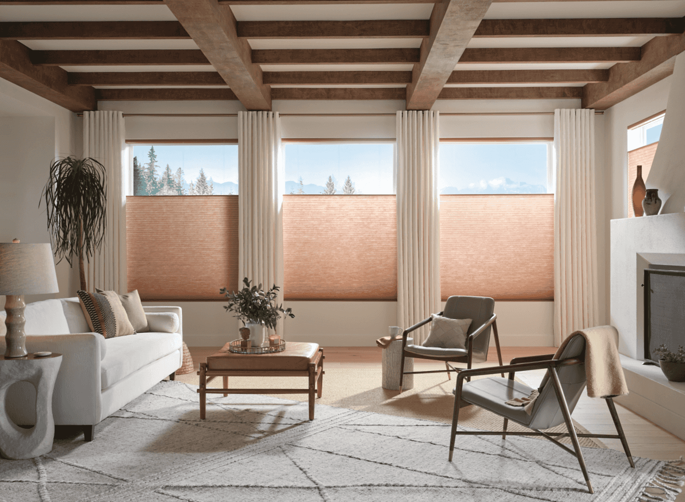 applause-sunterra-cornucopia-honeycomb-shades-living-room-wide Wide living room with three Applause Sunterra Cornucopia honeycomb shades in terracotta partially raised showing mountain views, with exposed wood beam ceiling and linen curtains