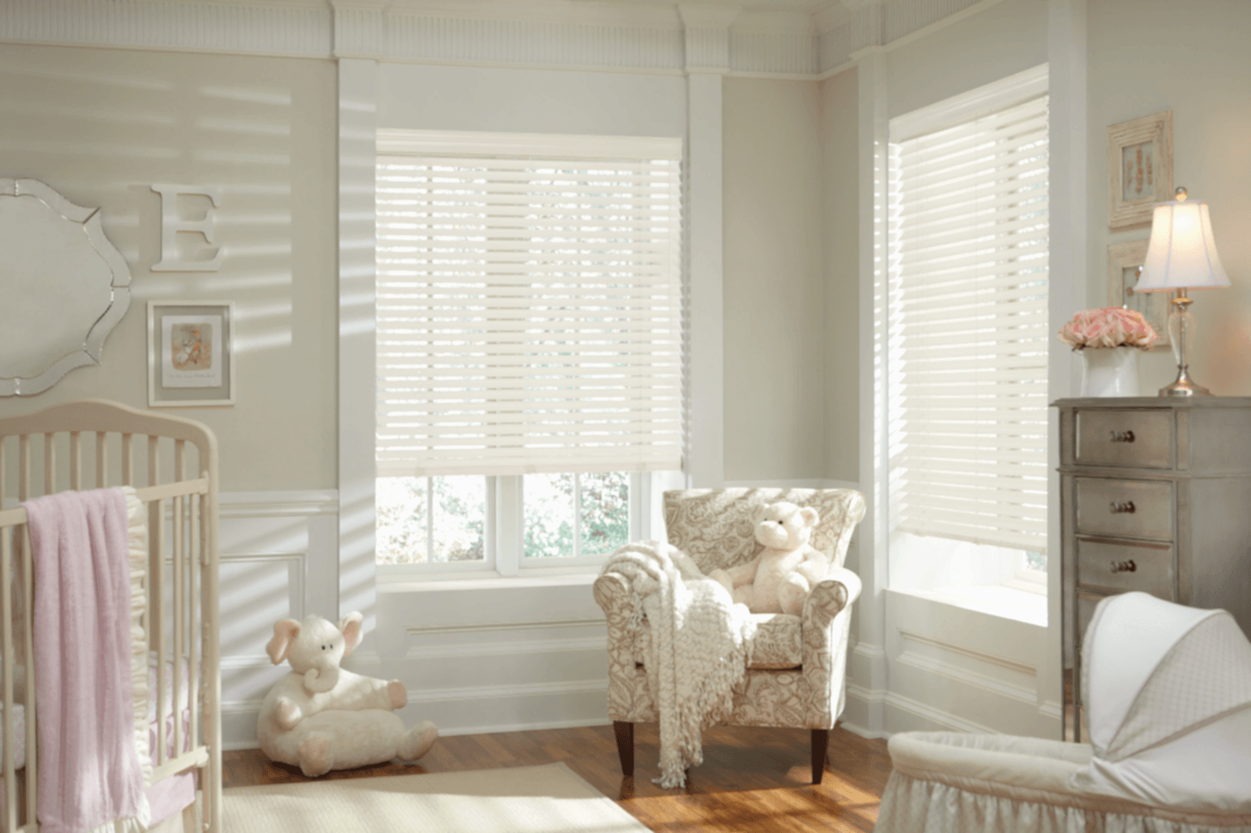 White wood blinds on double bay windows in a soft pink baby nursery, providing gentle light control and privacy for a child's room