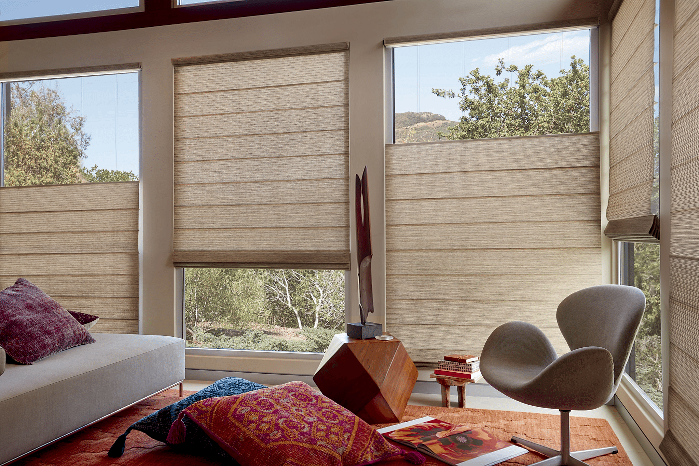 Natural woven roman shades in warm tan with visible horizontal fold lines installed on three windows in a sun-filled living room with colorful bohemian decor and hillside views